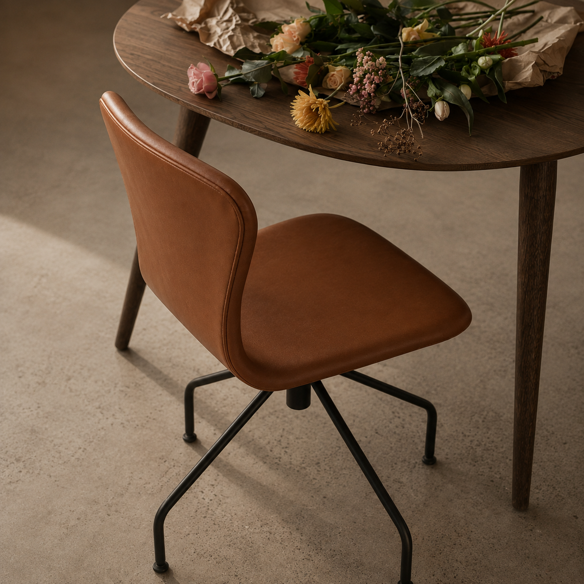A brown leather swivel dining chair with a black metal five-star base, positioned next to a dark wood table decorated with dried flowers on a textured concrete floor.
