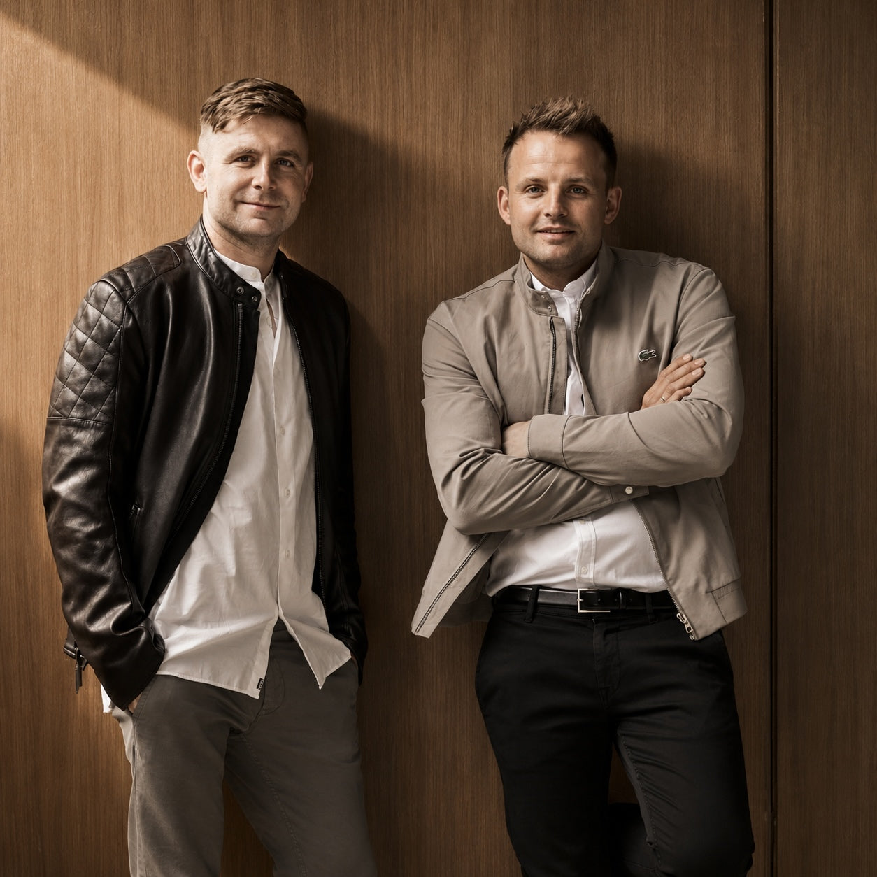 Two men standing against a wood-paneled wall, wearing casual jackets and shirts. The man on the left wears a black quilted leather jacket over a white shirt and gray trousers.