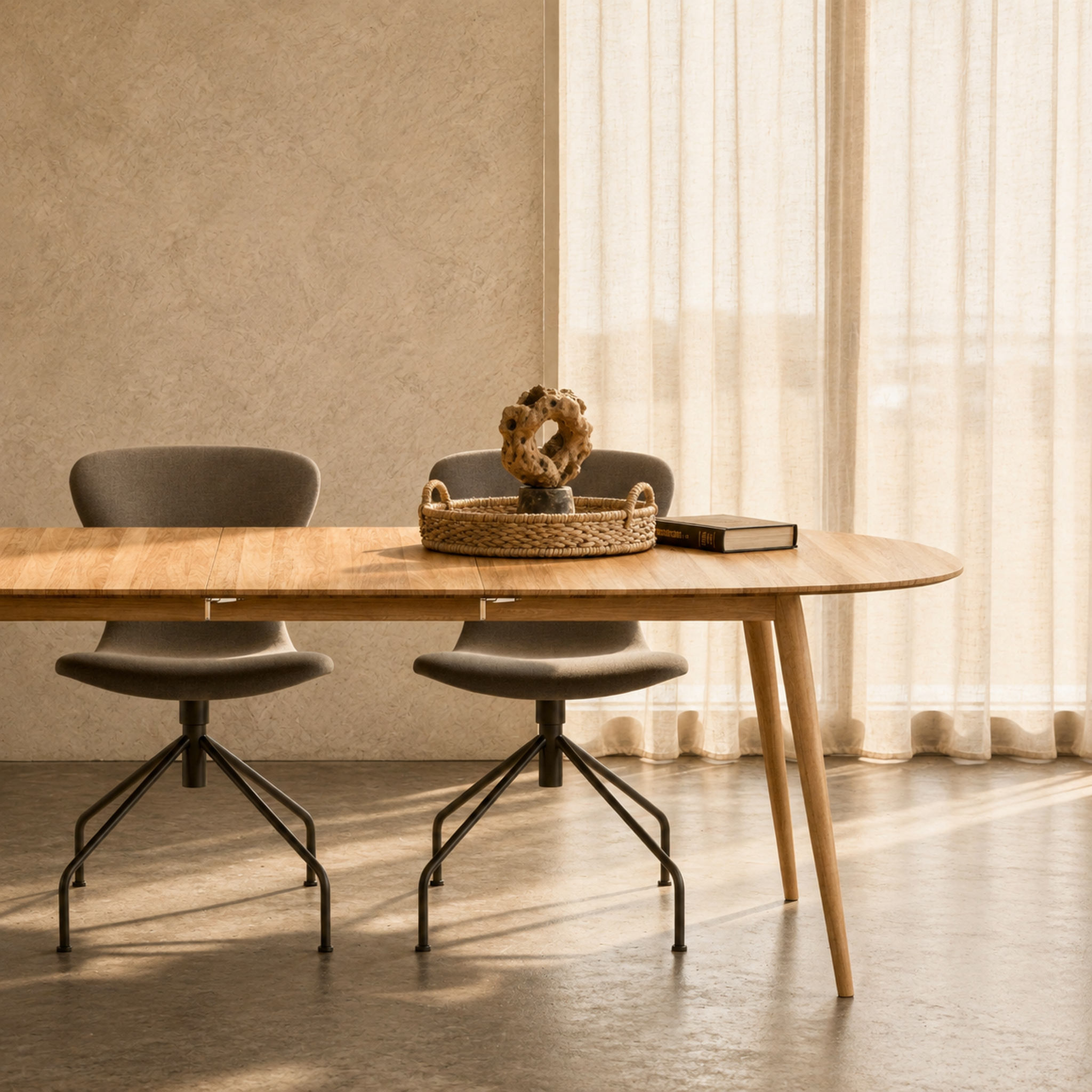 A modern oval wooden dining table with tapered legs, paired with two gray upholstered swivel chairs with black metal bases, set in a warm, sunlit room with textured beige walls and sheer curtains.